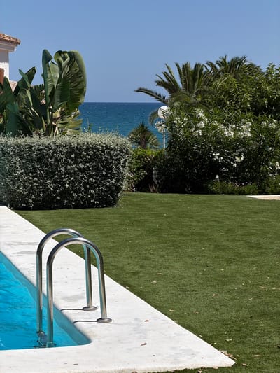 This is a vertical, eye-level, full shot of a swimming pool and its surrounding area on a sunny day, likely in a coastal region of Alcalà de Xivert, Spain, given the clear blue sky and ocean in the background. The foreground features the edge of a light-colored, concrete pool deck bordering a bright blue swimming pool. A silver metal pool ladder with two arched handles is partially submerged in the water. The pool deck transitions to a vibrant green artificial turf lawn that covers the majority of the mid-ground and extends to the right of the frame. In the background, a neatly trimmed, dark green hedge forms a boundary on the left, behind which are large, lush green leaves of tropical plants, and part of a white building with a tiled roof. To the right of the hedge and slightly further back, dense green foliage, including palm trees and flowering bushes with white blooms, create a layered backdrop. Beyond the vegetation, the deep blue ocean meets the clear blue sky at the horizon. The scene is devoid of any people or animals.