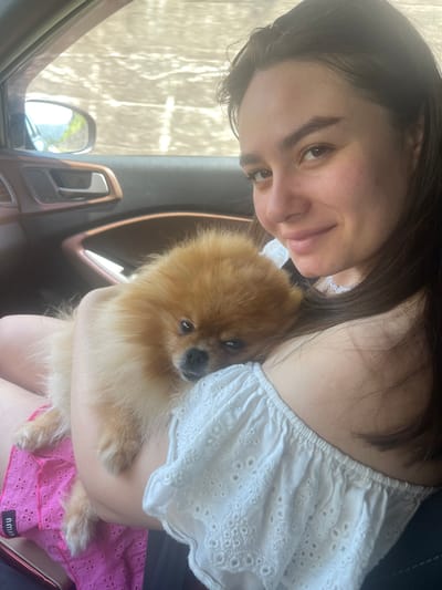 A young woman with dark hair is inside a car, holding a small, fluffy Pomeranian dog. She is smiling and looking towards the camera. The dog is nestled against her shoulder. The woman is wearing a white off-the-shoulder top with eyelet details and pink shorts. The car's interior is visible, including the door handle and window. The exterior view through the window shows a bright, slightly blurred background, suggesting movement. The location is identified as Simitli, Bulgaria.