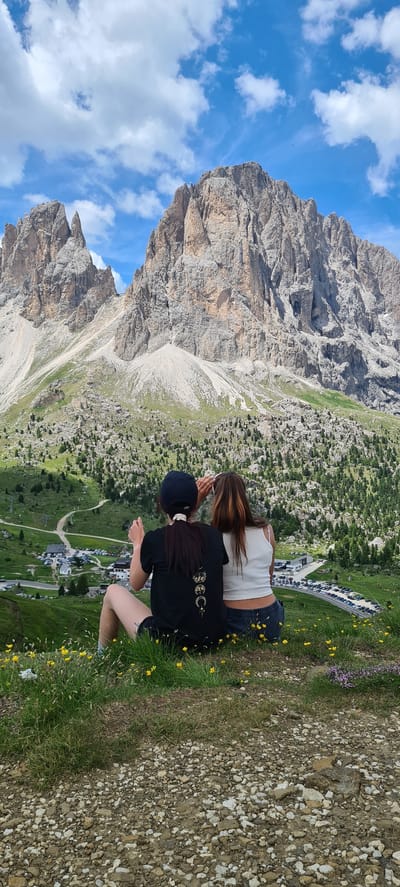 Two women sit facing away from the camera, looking out at a vast mountainous landscape. The woman on the left wears a black t-shirt with a graphic design and a black baseball cap. The woman on the right wears a white tank top. They are seated on a grassy hill dotted with yellow wildflowers. Below them, a winding road leads to a parking lot filled with cars, and several buildings. The background is dominated by massive, rugged mountains with steep, rocky slopes, partially covered in green vegetation and trees. The sky is blue with scattered white clouds. The location is in Val Gardena, South Tyrol, Italy, likely near the Sella Pass, given the distinctive mountain formations.