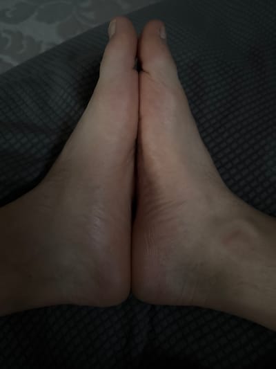 The image is a close-up, top-down view of two bare human feet, side-by-side, resting on a dark gray textured surface. The feet appear to be pale with visible veins and fine hairs on the skin. The toes are aligned, with the big toes touching. The lighting creates shadows that emphasize the contours of the feet and the texture of the surface. The setting is indoors and the surface appears to be a blanket or fabric. There is no indication of the location being in Sant'Angelo di Piove di Sacco, Italy.
