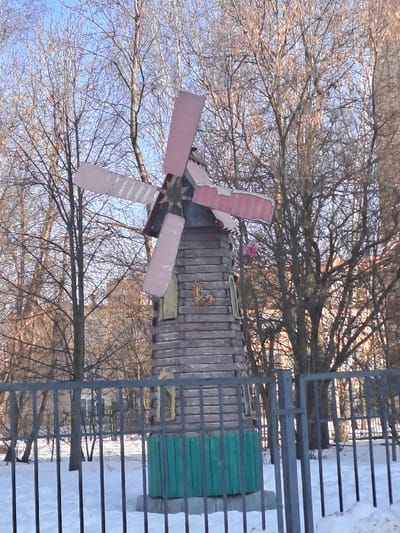 This forensic description details a still image captured in Yegoryevsk, Russia, depicting a decorative wooden windmill structure during daytime in a snowy, winter environment.

The primary subject is a freestanding, conical-tapered wooden windmill approximately 2.5 to 3 meters in height. Its lower section, roughly one-fifth of its total height, is painted a deep teal-green, featuring vertical planks. The main body of the windmill is constructed from horizontal wooden planks, painted a weathered grey-brown, exhibiting several small, rectangular, light-colored window-like cutouts or painted shapes. A golden-colored rooster or bird emblem is affixed to the upper part of the windmill's grey-brown body. The top of the structure features a dark, conical roof from which four broad, rectangular blades extend. These blades are painted in an alternating pattern of light pink/beige and dark pink/red and appear stationary. A small, spherical pink object, possibly a decoration, is visible attached to the right side of the windmill structure.

The windmill stands on ground completely covered in white snow. In the immediate foreground, a dark metal fence with vertical bars and two horizontal rails runs across the lower portion of the frame, partially obscuring the base of the windmill and the snow. The fence includes a gate section on the right side. The background consists of a dense arrangement of numerous bare deciduous trees, their branches devoid of leaves, indicating a winter or early spring season. Faint outlines of multi-story buildings are discernible through the bare branches in the distant background. The sky above the trees is clear and light blue. There are no individuals, active movements, or discernible interactions depicted in the scene.