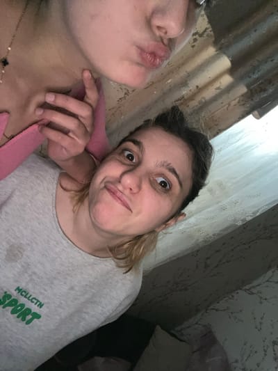 The image depicts two individuals in a close-up, tilted indoor setting. In the foreground, a person with light brown hair, appearing to be pulled back, looks directly at the camera with wide eyes and a slight smirk, their lips slightly pursed. This individual wears a light grey short-sleeved t-shirt with a visible dark stain on the upper left chest and green text that reads "MCLLCTN SPORT." Partially visible in the upper background is the chin, mouth, and neck of a second individual, making a "kiss" face with pursed lips. This person wears a pink top and a gold chain necklace featuring a small, black, four-leaf clover-shaped pendant. A hand, likely belonging to the second individual, is seen resting on the foreground person's left shoulder and neck area. The background features a corrugated or ridged light-brown surface, possibly a ceiling or wall, above the individuals. To the right, a white, semi-sheer curtain with a patterned design is visible, extending down to reveal a light-colored patterned wallpaper and a light-colored pillow or cushion. The environment appears to be an interior room. The specific location within the city of Lanchkhuti, Georgia, is not discernible from the visual evidence presented.