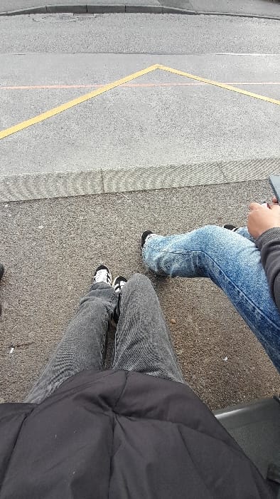 The scene, located in Souffelweyersheim, France, captures a downward perspective focused on the lower bodies of two individuals positioned on an asphalt surface adjacent to a roadway. In the immediate foreground, a person wears a black padded jacket, dark gray denim trousers, and black-and-white sneakers with black laces. To their right, a second individual is visible, wearing light blue acid-wash denim trousers and dark shoes with visible white soles or accents, possibly striped socks near the ankle. This second individual's right hand, clad in a gray long-sleeved garment, holds a dark, rectangular electronic device.
The ground beneath the individuals is textured dark gray asphalt. This area is bordered by a light gray, textured concrete curb or tactile paving strip, which separates it from a darker asphalt roadway beyond. Yellow lines forming a prominent inverted "V" shape are painted on the roadway surface, with a faint reddish-orange line running horizontally beneath them.