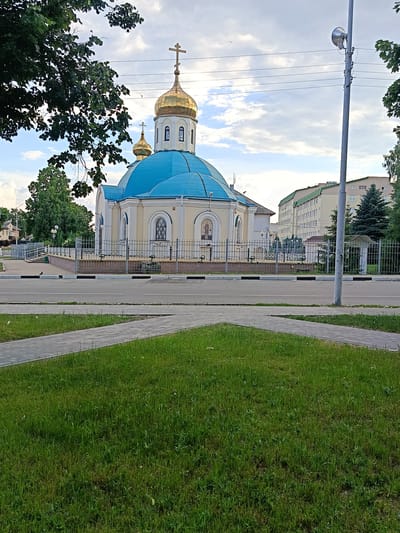 The image shows a medium-sized church with a prominent blue dome and a smaller golden dome, both topped with crosses. The church is painted a light yellow color with white trim. It is surrounded by a metal fence. In front of the church, there is a road, a sidewalk, and a grassy area with a pathway made of paving stones. To the right of the church, there is a tall lamppost with a speaker attached. In the background, there are several multi-story buildings and trees. The sky is partly cloudy. The scene is located in Gubkin, Russia, as indicated by a sign visible on the church. No people are visible in the image.