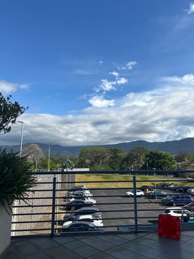The scene is overlooking a parking lot in Pozos, Costa Rica (latitude: 9.957, longitude: -84.196). In the foreground, there is a gray metal railing separating the parking lot from a cement walkway. A red traffic cone sits on the walkway. There are a number of parked cars in the lot, most of which are white and black. The parking lot is surrounded by a stone wall, which is overgrown with vegetation. In the background, there are mountains and trees. The sky is blue with white clouds. The view is from a parking garage with a street lamp to the left.