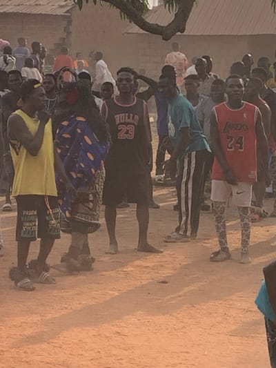 A group of people stand close together on a sandy surface outdoors. Several wear basketball jerseys; Lakers #24, Bulls #23, and others. Some individuals wear patterned clothing and athletic shorts. They are mostly Black. The overall scene is a dusty, possibly rural environment with a building on the left. The orientation of the photograph is rotated counterclockwise.