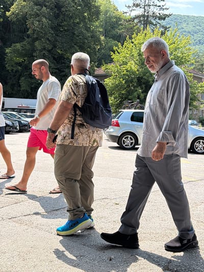 Three men are walking on a paved surface, possibly a parking lot, in Jajce, Bosnia and Herzegovina. The scene is outdoors with lush green trees and a hillside in the background. The time of day appears to be late afternoon, given the long shadows cast by the individuals. The weather seems warm and sunny.
The man in the foreground on the right is wearing bright blue and yellow running shoes, olive green pants, and a camouflage patterned short-sleeved shirt with a black backpack. He has a shaved head and is looking down.
The man in the middle is wearing a white t-shirt and bright pink shorts. He is also looking down and appears to be walking. Visible text on his shoes reads "HOKA".
The man in the background, on the left, is wearing a light gray collared shirt and gray pants with black slip-on shoes. He has a gray beard and is looking down and to his left. Behind him, a silver car is parked.
The overall impression is a casual outing or stroll.