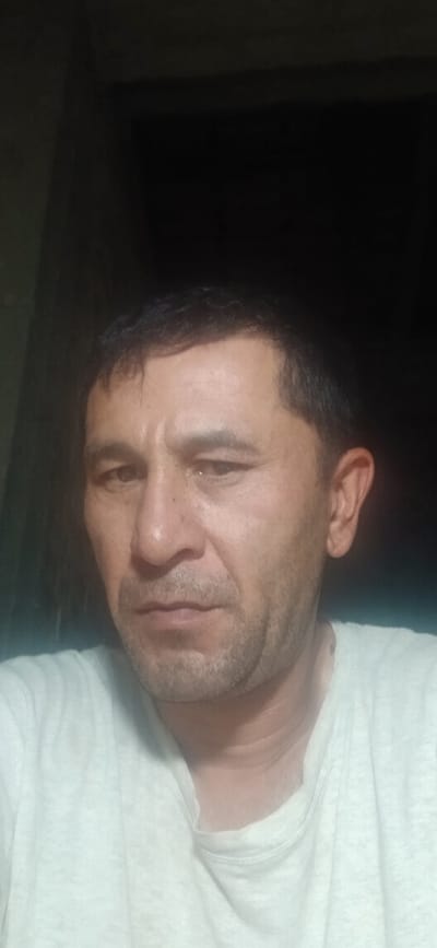 A man is pictured from the chest up, looking towards the camera. He has dark hair, brown eyes, and a light complexion. He is wearing a light-colored t-shirt. The background is dark and indistinct, suggesting an indoor setting at night or in low light conditions. There is no identifiable information about the location within Daliguzar, Uzbekistan, or any specific actions or interactions taking place.