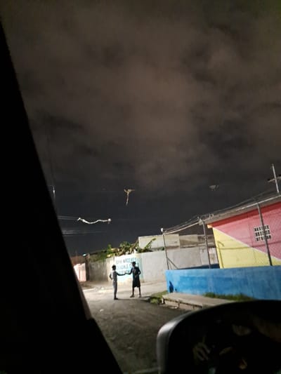 In the city of Cumana, Venezuela, at night, two young men are standing on a street in what appears to be a residential area. One man, dressed in a white t-shirt and light-colored pants, faces away from the camera and appears to be interacting with the other man, who is wearing a dark t-shirt and dark shorts. Both men are looking towards each other. In the background, there are low-rise buildings with painted facades in various colors, including pink and yellow. A chain-link fence separates the street from some of the buildings. The sky is dark and cloudy. Several kites are visible in the sky, including one long, serpentine kite and a smaller, more traditional kite. The scene suggests a casual, evening gathering or activity on the street.