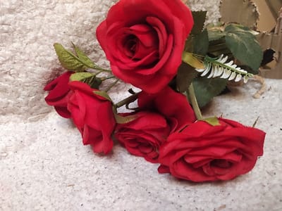 The scene is located in Harasta, Syria. The image displays a cluster of five artificial red roses resting on a light-colored, textured surface, possibly a carpet or fabric. One prominent red rose, appearing fully bloomed, is positioned at the top center. Below it, four additional smaller red roses or buds are visible, arranged in a semicircle. Each rose has artificial green leaves attached to its stem. A small, white, frond-like artificial floral accent is discernible among the green leaves near the upper rose. Brown twine is wrapped around the stems, visible towards the upper right. In the far upper right background, a piece of light brown corrugated cardboard with torn edges is partially visible. No people are present, and no actions or interactions are depicted. The lighting is even, suggesting an indoor setting.