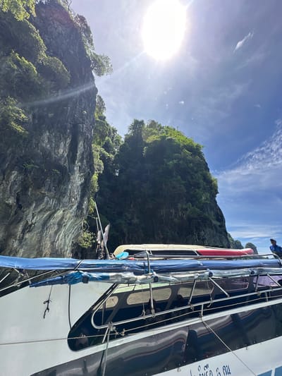 In Haad Hin Khom, Thailand, a boat is docked next to a sheer, rocky cliff face covered in lush green vegetation. The sun shines brightly from a clear blue sky with scattered clouds. The boat has a blue tarp covering part of its deck, and several kayaks or similar watercraft are strapped to the roof. A person wearing a blue shirt is visible on the boat, looking out towards the water. The rocky cliff is imposing, with trees clinging to its surface. The water around the boat appears calm.