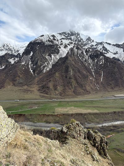 The image depicts a mountain landscape near Sioni, Georgia. A tall, snow-capped mountain dominates the left side of the frame. A road runs along the base of the mountain and through a grassy valley. A river flows parallel to the road. The foreground is a rocky cliff with dry grass. The sky is cloudy.
