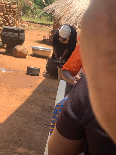 In Fada, Nigeria, the image shows an outdoor scene with a person wearing a black shirt and patterned blue and orange shorts sitting on a white chair. An individual wearing an orange shirt and a white head covering is bent over a large black pot. Next to the pot is a metal bowl, a piece of wood, and a large rock. The ground is brown dirt, and there is a thatched roof in the background. A stone wall and trees are visible in the distance.