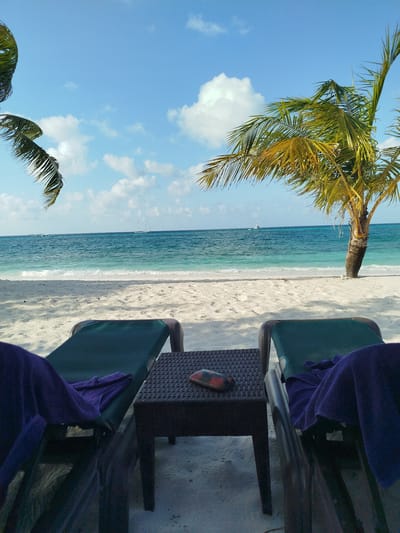 The image depicts a tranquil beach scene in Hinnavaru, Maldives. Two empty green lounge chairs are positioned on a white sandy beach, each draped with a purple towel. A dark, woven-patterned table sits between the chairs, holding a small, red and black object that appears to be an ashtray with a cigarette. In the background, the turquoise ocean meets the shore with gentle waves. A palm tree with green and yellow fronds stands to the right, casting a shadow on the sand. The sky is a clear blue, dotted with scattered white clouds. A sailboat and a small boat are visible in the distance on the water. The scene conveys a sense of relaxation and tropical serenity.