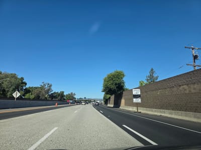 This image captures a perspective from inside a vehicle traveling on a highway in West Sacramento, California. The scene is set under a clear, bright blue sky, indicating a sunny day with good weather. The highway has multiple lanes, with other vehicles visible in the distance, suggesting moderate traffic.
To the right of the highway, a tall, textured brown brick wall lines the road, partially obscured by a large, leafy green tree. A signpost stands near the wall, displaying two white rectangular signs. The upper sign reads "SPEED LIMIT" and the lower sign states "RADAR ENFORCED."
On the left side of the highway, there is a concrete barrier with orange traffic cones visible in the distance, suggesting possible construction or maintenance. Beyond the barrier, a line of trees and foliage separates the highway from what appears to be a residential or commercial area. A white diamond-shaped sign with a reflective surface is also visible on the left.
In the far distance, an overpass with text that appears to read "WEST SACRAMENTO" can be seen, confirming the location. Overhead power lines and a utility pole are visible against the sky on the right side of the image. The overall impression is one of routine travel on a sunny day in a suburban highway environment.