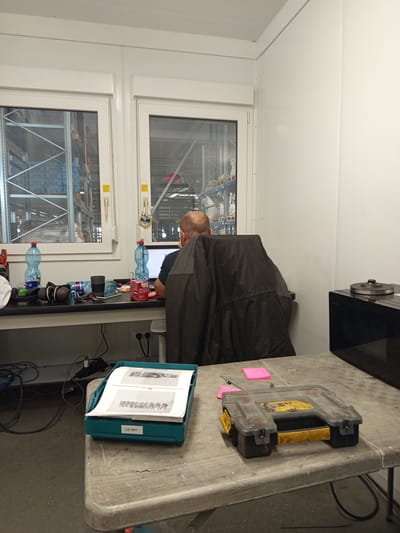 The image depicts an indoor scene, likely an office or workshop area, with a man seated at a desk in front of a computer. The desk is cluttered with various items, including water bottles, headphones, a cup, and a box of tissues. A dark jacket is draped over the back of the man's chair. Through a window behind the desk, a warehouse environment with shelving units and stored goods is visible. To the right of the desk, a black microwave oven is present. In the foreground, a grey table holds a teal bin containing documents and a yellow and black toolbox. The walls and ceiling appear to be made of white paneling. The overall impression is one of a functional workspace. The location of Vlkanová, Slovakia is not indicated in the image itself and would require external information to confirm.