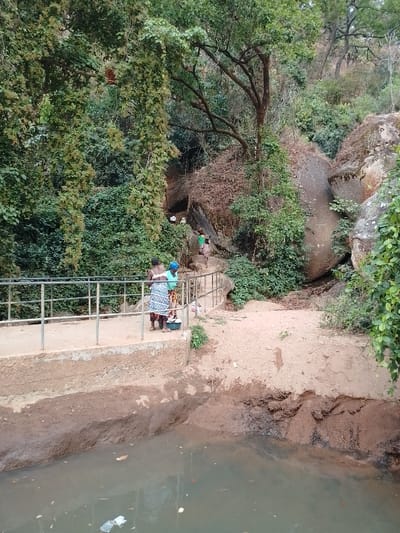 A scenic outdoor view features a body of water in the foreground, littered with trash. On the bank stands a concrete walkway with metal railings. Two women stand on the walkway. Behind them, a dirt path leads uphill through lush vegetation and massive rocks. Other individuals can be seen ascending the path. Dense foliage covers the hillside.
