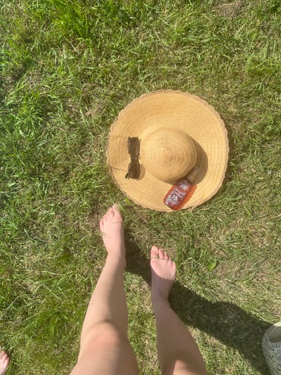A high-angle, overhead shot captures a sunny day on a grassy lawn. In the center of the frame lies a straw hat with a woven texture and a scalloped edge. Resting on the hat are a pair of dark-rimmed sunglasses and a bottle of Garnier Ambre Solaire with SPF 15. To the left of the hat, a pair of bare feet and legs are visible, extending upwards towards the top of the frame. The grass is green with patches of dry, brown blades, indicating a warm season. The sunlight casts a subtle shadow to the right of the feet, suggesting the sun is positioned to the left. The scene is set outdoors on a bright, clear day, likely in a park or backyard.