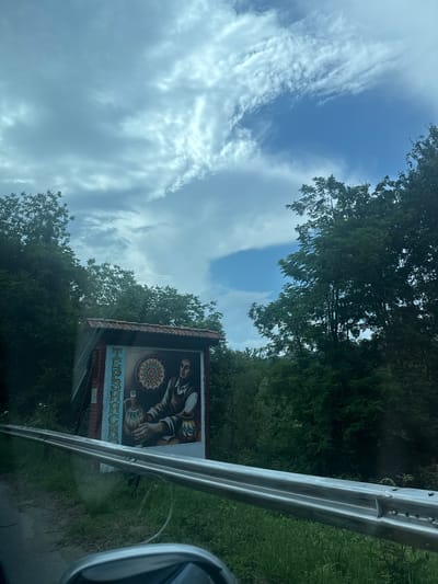 The image captures a roadside scene under a cloudy sky, likely in or near the Bulgarian city of Terziysko. In the foreground, a metal guardrail runs along the edge of a road, with a grassy verge to its right and a dirt shoulder to its left. The main subject is a large, colorful mural painted on a structure beside the road, under a red-tiled roof. The mural depicts a man in historical attire, holding two ornate jugs and standing in front of a circular, decorative design. Vertical text in a Cyrillic alphabet is also visible on the left side of the mural, and appears to spell out "Terziysko". Lush green trees and foliage form a dense backdrop, with a patch of bright blue sky peeking through the clouds. The perspective suggests the photo was taken from inside a vehicle.
