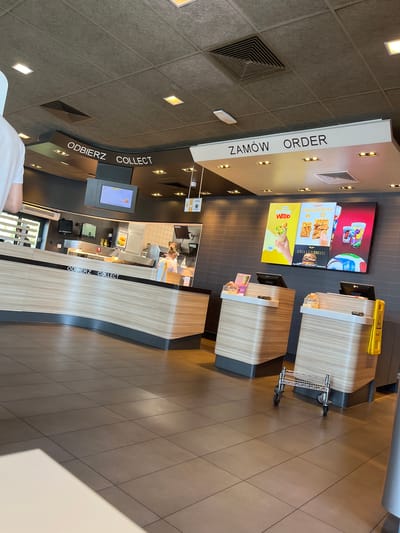 The image depicts the interior of a McDonald's restaurant in Katowice, Poland. The scene is viewed from the perspective of a customer looking towards the counter area.
The ceiling is a neutral gray, with recessed lighting fixtures and air vents visible. Along the ceiling, above the counter, are signs in Polish and English. To the left, the sign reads "ODBIERZ COLLECT," indicating a pickup point for orders. To the right, the sign reads "ZAMÓW ORDER," indicating the ordering area.
The counter itself is a light wood-grain laminate with a curved design. In front of the counter, there are two digital ordering kiosks, each with a touch screen display. On the wall above the ordering area, there are illuminated menu boards showcasing various food items, including wraps, chicken products, and drinks. One board prominently features a hand holding a wrap.
Behind the counter, the kitchen area is visible, with stainless steel surfaces and equipment. A female employee wearing a light-colored uniform is seen working behind the counter, with her back to the camera. She has blonde hair tied up.
The floor is tiled with large, dark gray square tiles. A small metal trolley with wheels is parked on the floor in the foreground, to the right of the ordering kiosks. A yellow safety sign is visible on a pillar near the trolley. The overall impression is of a modern and clean fast-food restaurant environment.
