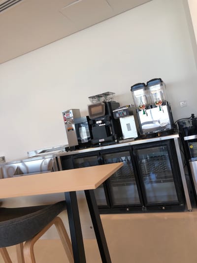 The image depicts a commercial self-service beverage and snack station located within a facility in Hanak, Saudi Arabia.
In the foreground, the corner of a light-colored rectangular table with a black metal base is visible, accompanied by a chair featuring a grey upholstered seat and wooden legs.
The midground features a stainless steel countertop setup against a plain, light-colored wall. From left to right on the counter: a stainless steel sink or preparation area, a silver-colored BUNN hot water dispenser, a black commercial coffee brewer, a black coffee grinder with a clear hopper containing coffee beans, another black coffee machine, and a silver-colored commercial beverage dispenser with two clear cylindrical containers, each filled with colorful, spherical candy-like items. Further to the right, a black countertop appliance, possibly another dispenser, is partially visible. Below the countertop, a three-door under-counter refrigerator unit with glass doors and black frames is present, displaying internal shelves. An additional black appliance stands to the right of the refrigerator unit.
The background consists of a plain, light-colored wall with a white electrical outlet. The ceiling is white with a visible rectangular vent and a flush access panel. No people, subjects, or actions are depicted in the scene. The overall environment appears clean and functional, indicative of a breakroom, cafeteria, or retail food service area. The image has a slight upward tilt from the lower-left to the upper-right.