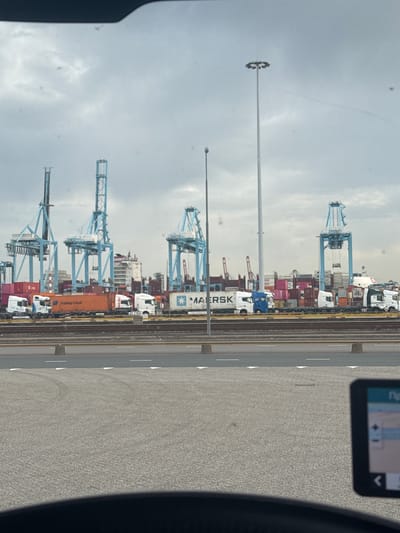 The image depicts a view from a vehicle, likely a car, in Maasvlakte Rotterdam, Netherlands. The scene shows a port area with several large gantry cranes against a cloudy sky. Container trucks are lined up along what appear to be train tracks or a loading area. A tall light pole is visible in the mid-ground. A navigation device is mounted on the car dashboard to the right. The road is covered in tire marks and shows wear.