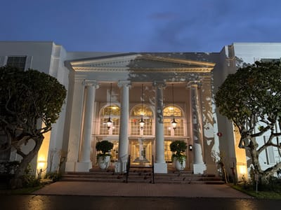The image depicts the entrance of a building at dusk or night. The architecture is neoclassical, featuring a portico supported by large white columns. Above the columns, there is a triangular pediment with decorative molding, and the facade of the building is illuminated by outdoor lighting, casting shadows from nearby trees onto the structure.
The entrance itself consists of a set of double doors with the number "10150" visible above them. Flanking the doors are large arched windows, also illuminated from within. Hanging lanterns with a classical design are suspended in front of these windows.
In front of the building, a set of wide steps made of brick leads up to the entrance. These steps are lined with manicured bushes and two large white planters, each containing a topiary-like plant.
On either side of the steps, mature trees with gnarled branches are visible. They are lit from below, highlighting their unique shapes and casting more shadows. The ground in front of the building is paved with brick, and to the right, there is a darker area, possibly a street or sidewalk, with reflections from the lighting, suggesting it might be wet.
The sky above is a deep blue, indicating twilight or night, with some faint clouds. The overall impression is one of elegance and grandeur, with the lighting accentuating the architectural features.
The building is located at 10150 Collins Avenue in Bal Harbour Village, Florida, United States of America.