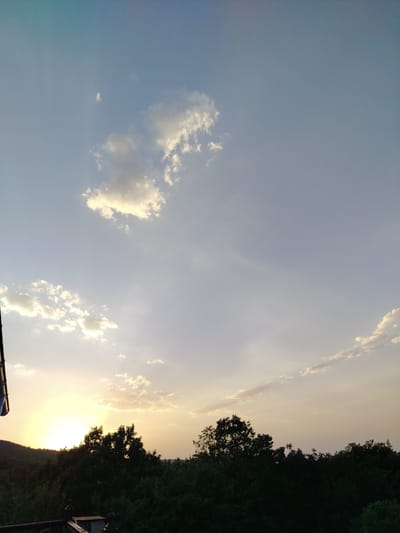 This is a photograph of a sunset over a forested area, likely in or near Galaalti, Azerbaijan. The sky is a gradient of blues and yellows, with scattered clouds catching the golden light of the setting sun. The sun itself is a bright, hazy orb on the horizon, casting rays through the atmosphere. A dark silhouette of dense trees forms the foreground, with a rolling hill visible behind them on the left. A dark railing or structure is also present in the lower portion of the image. The overall scene depicts a natural landscape at dusk.