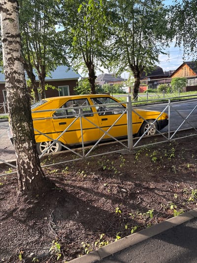 A yellow, four-door sedan is parked behind a metal fence. The car appears to be older, with visible wear. The scene is located in the city of Rezh, Russia, as indicated by the presence of typical Russian dacha-style houses and wooden fences in the background. The area around the car is covered in dirt and sparse vegetation, with a large tree on the left side of the frame. The sky is clear and blue, suggesting it is daytime. There are no people visible in the image. The lighting suggests it is late afternoon or early morning, with long shadows cast by the trees.