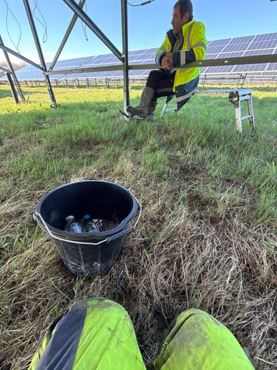 The image captures an outdoor scene at a solar farm located in Eastleigh, United Kingdom. The foreground features the lower torso and legs of an individual, wearing neon yellow and black work trousers exhibiting visible soiling. Adjacent to this, a black plastic bucket with a metal handle rests on the ground, containing two plastic water bottles (one clear, one with a blue cap) and dark cables or wires. The ground consists of a mixture of green and dry, brownish grass with patches of exposed soil.
In the midground, a male subject is seated on a metallic, foldable step stool positioned beneath the framework of a solar panel array. He wears a neon yellow and dark blue high-visibility work jacket, dark trousers, and dark, muddy work boots. His hands are clasped, resting on his lap, and he is looking towards the right side of the frame. The background reveals multiple rows of solar panels extending into the distance under a clear, bright blue sky, with their supporting metal structures clearly visible. A line of trees marks the horizon on the far left.