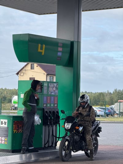 Two men are present at a green and grey fuel pump island located at a gas station in Starovoitove, Ukraine, on an overcast day.
To the left, a man stands beside the pump unit. He wears a dark grey hooded vest over a green long-sleeved shirt with a reflective stripe, and dark trousers. He holds a small, light object near his face and has a white plastic bag containing gloves attached to his left hip.
To the right, a second man is seated astride a black motorcycle. He is dressed in camouflage attire, including a jacket and trousers, and wears a white and black full-face helmet with a dark visor. He holds and is looking down at a smartphone with both hands. The black motorcycle has a front headlight, handlebars, and a visible exhaust pipe. A dark backpack is worn by the rider.
The fuel pump unit is predominantly green with a prominent yellow number "4" on its upper display. It offers multiple nozzles for "ДП" (Diesel) and "95" octane gasoline, labeled with "Mustang 95," "AVI Cappe 5," and "ДП Prime" on small screens. A poster with Ukrainian text is affixed to the pump. The base of the pump shows "B-1r" and hazard symbols.
In the background, behind the pump, a light-colored, possibly two-story, residential house with a dark brown roof and a security camera is visible. A line of green trees forms the horizon. To the far left, a portion of a white and green tanker truck is visible behind a fence. To the far right, various parked cars and a larger vehicle are discernible in a lot. The sky is cloudy.