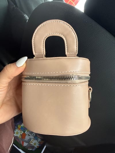 The scene depicts a light beige cylindrical handbag with a semi-circular top handle and a metallic zipper, held by a person's hand with white-painted fingernails. The handbag is positioned against a dark, textured background consistent with vehicle upholstery. Partially visible below the handbag is a colorful, blurred item featuring cartoon characters, including what resembles Bugs Bunny amidst carrots and daisies. A red and white object is subtly visible in the upper right background. The event is located in the city of Apnagyugh, Armenia.