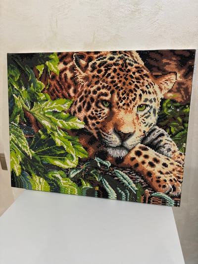 The image shows a diamond painting of a jaguar nestled amongst green leaves. The painting is positioned horizontally and is framed by a black border. The jaguar's face is the focal point, with its green eyes and spotted fur rendered in intricate detail using small, colored gems. The leaves surrounding the jaguar are depicted in various shades of green, with some lighter green highlights suggesting sunlight or dew. The background is dark, creating a sense of depth and emphasizing the jaguar and foliage. The painting rests on a white surface, likely a table or shelf, and appears to be displayed against a textured, light-colored wall. There is no indication of the city of Ozerne, Russia, within the image itself.