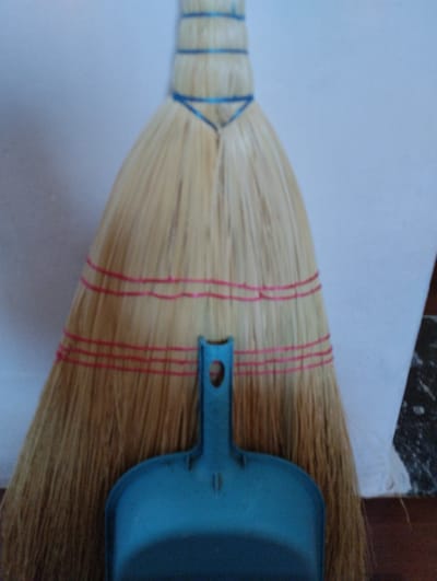 A close-up, overhead shot shows a straw broom with a blue plastic dustpan positioned underneath the bristles. The broom is bound with blue string near the top and has three horizontal rows of red string wrapped around the bristles further down. The dustpan is a solid blue color and has a curved lip. The background consists of a light-colored wall on the left and a white sheet or cloth on the right, with a dark patterned surface visible at the bottom right. The lighting is soft and diffused. The image does not provide any information or visual cues about the city of Yozgat, Turkey.