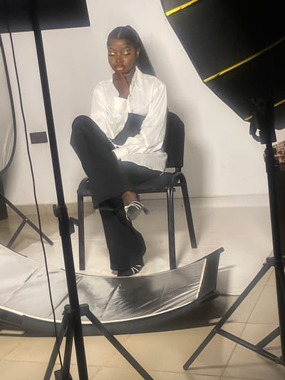 A young woman with dark skin and long dark hair pulled back in a ponytail is seated on a black metal chair in what appears to be a studio setting. She is wearing a loose-fitting white button-down shirt tucked into high-waisted black pants. Her makeup includes bold gold eyeshadow and bright pink lipstick. Her left hand is resting on her chin, and her eyes are looking down. Her feet are crossed, and she is wearing black and white striped sandals.
The background is a plain white wall. To the left and right of the woman are large studio lights. In front of the woman, on the floor, is a large, curved reflector. The floor is tiled with light-colored tiles. A power outlet is visible on the white wall behind the woman.
The image does not provide any information about the specific location in South Ropp, Nigeria.