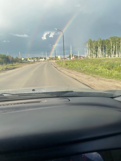 The image is taken from the driver's perspective inside a vehicle, looking out at a road. A double rainbow is visible in the overcast sky. The road appears to be a two-lane asphalt surface leading into the distance, with a grassy verge and a line of birch trees on the right side of the road. In the distance, there are some buildings and streetlights. The foreground shows the dashboard of the vehicle. The scene is consistent with a suburban or rural area. The location is identified as Prigorodny, Russia.