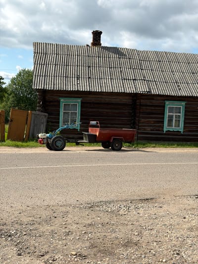 In Byahoml, Belarus, a scene unfolds before a weathered wooden building. The building's dark logs contrast with the corrugated, slate-grey roof, capped by a brick chimney. Two windows, framed in vibrant turquoise trim, punctuate the facade. To the left of the building, two tall, orange-yellow doors stand leaning against the exterior, accompanied by a weathered grey wooden panel.
In front of the building, a small, blue and white motorized cultivator sits parked, its red engine housing and larger rear wheels prominent. It is hitched to a red, two-wheeled trailer. The trailer is empty, with visible text on its side. The cultivator and trailer rest on a patch of grass and dirt at the edge of an asphalt road, which occupies the foreground of the image. The road is marked with a faint white line. The ground bordering the road is a mixture of gravel and dirt, with small stones scattered across it. The sky above is a mix of blue patches and heavy grey clouds, hinting at changeable weather. Trees are visible in the background, to the left of the building. The overall impression is one of a rural, utilitarian setting.
