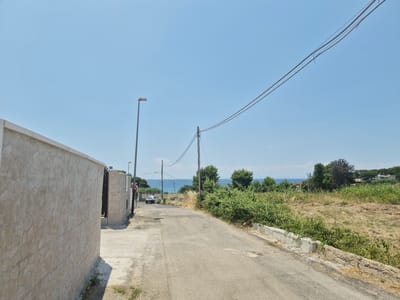 The image captures a view of a paved road leading towards the coast in Leporano, Italy. On the left, a light-colored stone wall runs along the side of the road. Further down the road, a person stands near an entrance. The road curves slightly and is bordered by dry vegetation and green bushes. In the distance, the blue sea is visible under a clear sky, with trees and some buildings on the horizon. Power lines stretch across the sky, supported by utility poles. A white car is parked further up the road. The scene appears to be a quiet, rural coastal area.