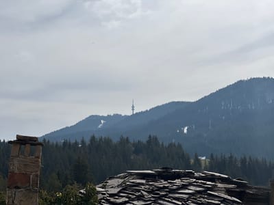 The image presents a view of a mountainous landscape in Stoikite, Bulgaria. In the foreground, there is a stone chimney to the left and a roof covered in dark gray slate tiles to the right. Behind them, a dense forest of evergreen trees covers the slopes of the mountains. In the middle distance, a tall, thin tower-like structure stands on the ridge of a mountain. Patches of snow are visible on the mountain slopes. The sky is overcast, with a layer of gray clouds. Power lines can be seen running through the forest. The general atmosphere is hazy and muted.
