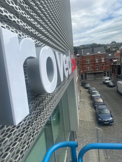 The image is an outdoor, high-angle shot of a street scene in Liège, Belgium. Dominating the foreground are large, white, three-dimensional letters spelling out "MOVENPICK", mounted on a perforated metal facade. The letters are sleek and modern, with rounded edges. Behind the facade, a line of cars is parked along a cobblestone street. The cars are a mix of dark blue, silver, and white. Further back, red-brick buildings with multiple windows and pitched roofs line the street, suggesting an urban residential or commercial area. The sky is visible in the upper left corner, filled with dramatic, streaky white clouds against a light blue background, indicating a partly cloudy day. A bright blue pipe structure is visible on the right side of the image, partially obscuring the view of the street and buildings, and appears to be part of nearby construction or infrastructure. The scene is brightly lit, suggesting it's daytime, likely mid-morning or afternoon given the angle of the light and shadows. There is no visible activity of people in the frame.