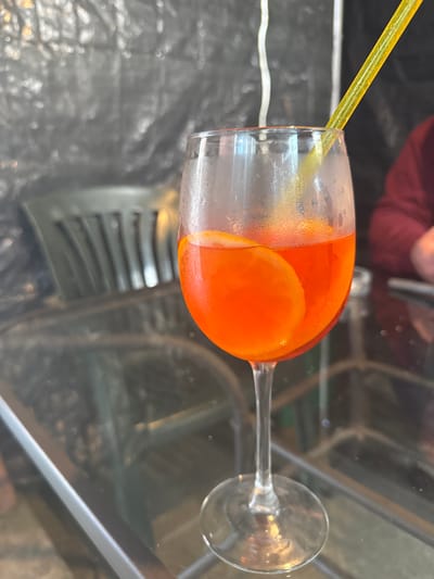 The image displays a close-up, eye-level perspective of a cold beverage served in a wine glass on a reflective glass table in Boston, United Kingdom. The glass contains a vibrant orange liquid, consistent with a spritz cocktail, and is garnished with a translucent orange slice submerged within. Condensation is visibly adhering to the exterior surface of the glass. A yellow, textured plastic straw is inserted into the drink, angled upwards and to the right from the glass rim. The background is significantly out of focus, featuring blurred reflections and shapes. To the left, a dark grey or black slatted chair is partially visible. The upper background shows a textured, possibly metallic or reflective dark surface with irregular light reflections. To the right, a segment of a person's upper arm or shoulder, covered in a maroon fabric, is discernable, also out of focus. The overall lighting suggests an indoor or covered outdoor setting with ambient illumination.