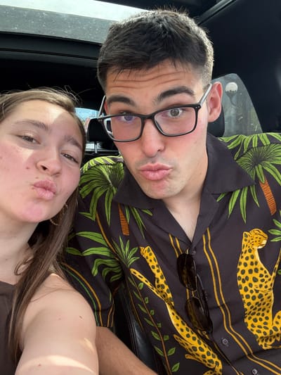 The image, taken in Radinovo, Bulgaria, captures a close-up selfie of two individuals, a male and a female, within what appears to be the interior of a vehicle. The perspective is from a low angle, looking upwards at the subjects, who are positioned close together in a playful pose. Both have their lips slightly puckered.
The male subject, prominent in the upper and central part of the frame, wears black-framed glasses and has short, dark hair. He is dressed in a black shirt adorned with a distinctive pattern of green palm leaves and yellow cheetah or leopard figures. A pair of dark sunglasses is casually tucked into the collar of his shirt.
The female subject is in the lower section of the frame, featuring long brown hair and wearing a dark, possibly brown, top. Her expression mirrors the male's, contributing to the casual and lighthearted mood of the photograph.
The scene is brightly illuminated by natural daylight, suggesting the picture was taken during daytime, likely under sunny conditions, as indicated by the clear reflections in the male's glasses and the general brightness. No discernible text is visible on clothing, objects, or in the background. The overall impression is one of a shared, affectionate moment.