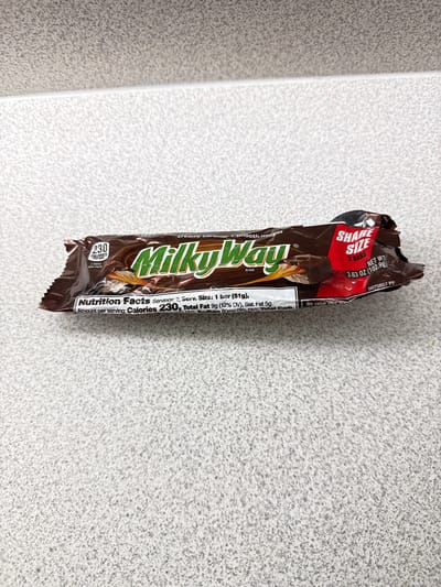 A Milky Way candy bar wrapper is pictured lying flat on a speckled gray and white surface. The wrapper is mostly brown with green lettering that spells out "Milky Way". There are also nutritional facts printed on the wrapper. The wrapper appears slightly crumpled.