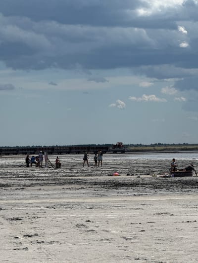 The image depicts a wide, flat expanse of salt flats, likely located in Bursol, Russia, under a dramatic, overcast sky filled with dark, heavy clouds. In the distance, a long train of rail cars stretches across the horizon, with a red locomotive visible on the right side. Several scattered groups of people are present on the salt flats. In the left middle ground, a group of four individuals are gathered, with one person appearing to paint or draw on an easel. Further to the right, a group of three people are visible, and in the far right middle ground, two individuals are relaxing on what appears to be a large tire. Scattered on the salt flats are various personal items including shoes and bags. The ground itself is a textured mix of light-colored salt and darker, muddy patches, suggesting a recently dried-up body of water.