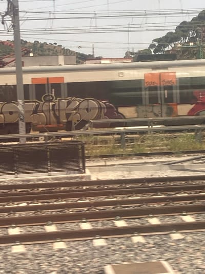 A train with graffiti on its side is seen traveling on railroad tracks in Montcada i Reixac, Spain. The train cars are light-colored with orange rectangular sections. The graffiti is elaborate and covers a significant portion of the train's side, featuring stylized lettering in various colors, including beige, black, and red. In the background, there are rolling hills covered with vegetation and trees, and overhead power lines suggest an active railway line. The foreground shows multiple sets of parallel railroad tracks with ballast.