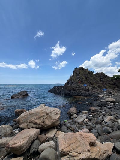 The image shows a rocky coastline with the sea and a bright blue sky. In the foreground, there are many large and small rocks of various colors, including tan, gray, and dark gray. Some of these rocks are smooth and rounded, while others are rough and jagged. The sea is a deep blue color, with gentle waves lapping at the shore. In the middle ground, there is a large, dark rock formation that juts out into the sea. This formation is covered in vegetation and has a path leading up to it, marked by a rope fence and several blue signs. In the background, the sea stretches to the horizon, meeting the sky under a partly cloudy sky with scattered white clouds. The overall scene is a natural landscape with a sense of rugged beauty. This location is likely a coastal area in Phú Lương, Vietnam, possibly a scenic spot or a nature reserve.