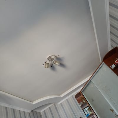 The image shows an indoor scene, likely a room, with a white ceiling that has decorative molding. A multi-light fixture is centrally mounted on the ceiling. To the right of the image, a portion of a wooden wardrobe with sliding glass doors is visible. The doors have a frosted floral pattern. A small shelf unit is attached to the left side of the wardrobe, containing a few decorative items and a framed picture. The walls are decorated with striped wallpaper in shades of grey and white. There is no indication of the specific location within Ovidiopol, Ukraine.