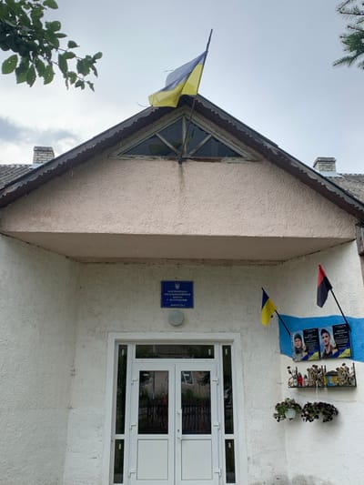 The image captures the front entrance of a building identified as "Lazeshchyna General Educational I-III Degrees, Building #2" in Lazeshchyna, Ukraine. The structure features a textured, light-colored stucco facade and a gabled roof with dark, scalloped trim and an integrated triangular window. A tattered Ukrainian flag (blue over yellow) is affixed to the peak of the gable.

The main entrance consists of modern white double doors with glass panels, recessed under an overhang. Above the doors, a blue rectangular sign displays the Ukrainian coat of arms and the aforementioned building identification in Cyrillic script.

To the right of the entrance, mounted on a wall section painted with blue and white horizontal stripes, are two smaller flags: a Ukrainian flag on the left and a red and black flag on the right. Below these flags, two memorial plaques display portraits of individuals, one female and one male, both appearing in military attire. Beneath the plaques, a wrought-iron shelf holds multiple miniature Ukrainian flags, several red objects resembling candles or votives, and other small, indistinguishable decorative items. Two hanging baskets containing dark-flowered plants are suspended below the shelf.

The sky overhead is overcast. Tree branches with green foliage are partially visible in the upper left corner of the frame. No individuals are present, and no actions or interactions are depicted.
