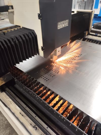 The image captures a close-up view of a laser cutting machine in operation. The machine is actively cutting a sheet of metal, evidenced by the bright orange sparks erupting from the point of contact. The laser head, a dark rectangular component with the inscription "ISMÖYİL" and "MAGNETENERJİ," is positioned above the metal sheet. The sheet itself is large and appears to be a work-in-progress, with several rows of circular cutouts already visible. Below the metal sheet, a system of sharp, interlocking metal supports is visible, designed to hold the material during the cutting process. These supports are coated in a dark, gritty residue, suggesting extensive use. The background of the image is slightly blurred but shows industrial machinery and equipment, including a pleated black bellows system on the left side of the frame, likely part of the machine's enclosure. The overall environment appears to be a workshop or factory floor. The image does not provide any visual information that would allow for the specific location in the city of Dangir, Uzbekistan to be identified.