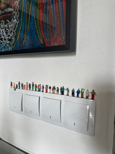 A line of miniature figures stands on a white wall-mounted light switch plate in Ammeldingen bei Neuerburg, Germany. The figures, varying in size and color, are arranged from left to right. They appear to be a diverse group, including adults and children, some dressed in business attire and others in casual clothing. Behind the light switch plate, a textured white wall is visible. Above the light switch plate, a framed piece of art with abstract, colorful lines in red, blue, yellow, and white is mounted on the wall. The artwork has a black frame. The scene suggests an indoor setting, possibly a residential or public space, with a creative arrangement of decorative elements.